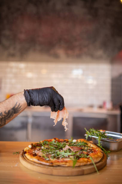 Unrecognizable chef, with protective gloves on, holding the slice of prosciutto above the pizza